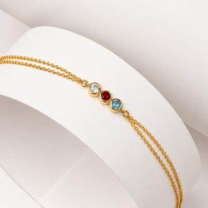 Può includere: Un braccialetto a doppia catena color oro con tre pietre preziose rotonde incastonate a lunetta in fila. Le pietre includono una gemma bianca trasparente, una gemma color rosso granato e una gemma color blu brillante in montature dorate.