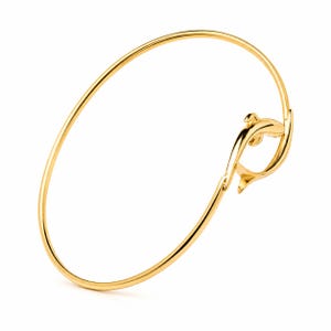 Op de afbeelding: Een gepolijste goudkleurige armband met een slank, gestileerd dolfijnmotief dat in de cirkelvormige band is geïntegreerd. Het metalen oppervlak is glad en reflecterend en vormt een doorlopende, elegante lus die naar de dolfijn toe buigt.