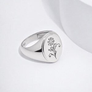 May include: A polished silver signet ring featuring an oval face with an engraved floral motif. The interior of the band is clearly stamped with the text 925, indicating the metal composition. The ring has a smooth, reflective finish throughout.