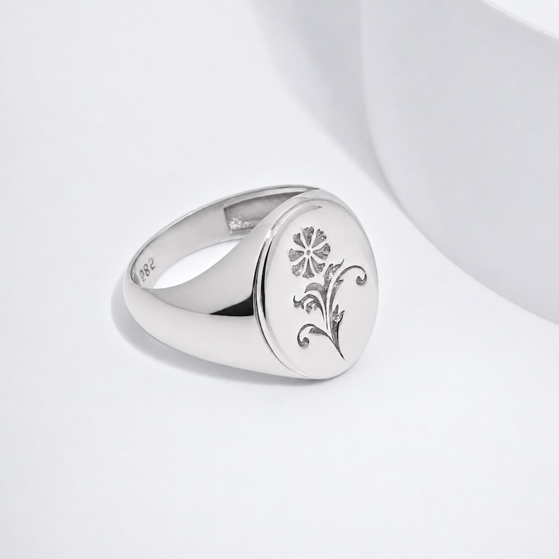 May include: A polished silver signet ring featuring an oval face with an engraved floral motif. The interior of the band is clearly stamped with the text 925, indicating the metal composition. The ring has a smooth, reflective finish throughout.