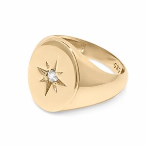 May include: A polished gold signet ring featuring a round face with a starburst engraving at the center. A small, clear, round-cut gemstone is set in the middle of the star. The interior of the band is clearly stamped with the hallmark 585.