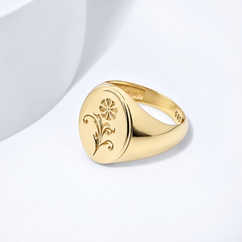 May include: A polished gold signet ring featuring an oval face with an engraved floral motif. The interior of the band is clearly stamped with the hallmark 585, indicating the gold purity of the piece. The ring has a smooth, reflective finish.