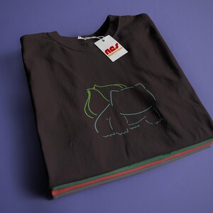 May include: A black t-shirt featuring a green and white outline of a cartoon creature. The shirt is folded and stacked on top of other shirts in different colours.