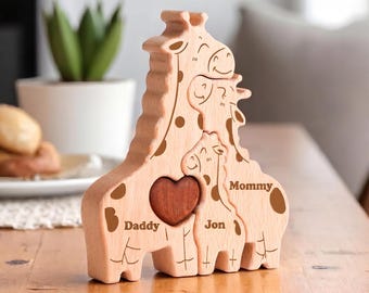 May include: A light-colored wooden puzzle shaped like three giraffes standing together. The puzzle features engraved text reading Daddy, Mommy, and Jon, with a dark brown wooden heart shape embedded in the center of the largest giraffe figure.