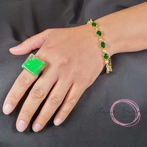 May include: A green square ring with a clear resin design. The ring is on a person's finger. The person is also wearing a gold bracelet with green oval stones.