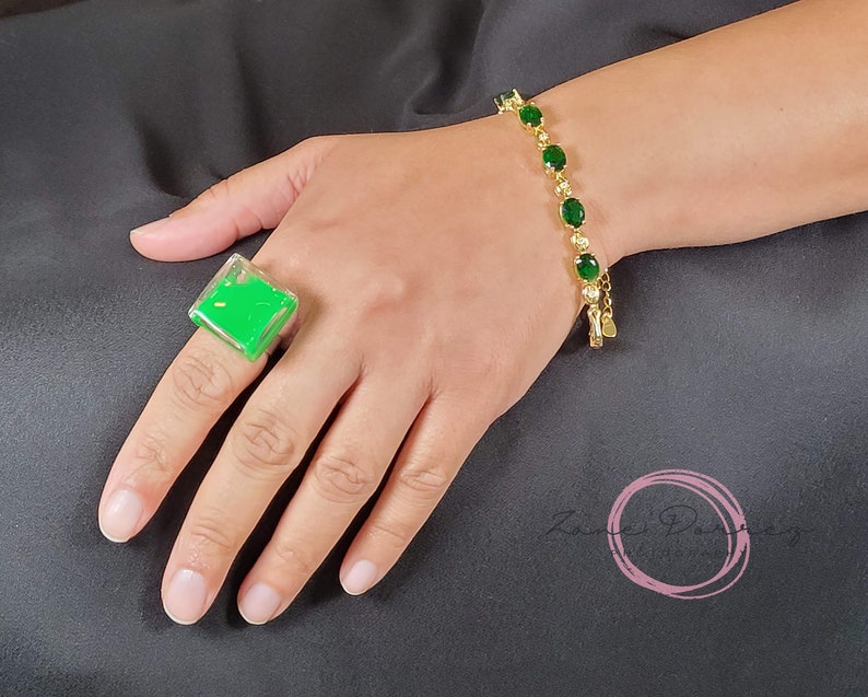 May include: A green square ring with a clear resin design. The ring is on a person's finger. The person is also wearing a gold bracelet with green oval stones.