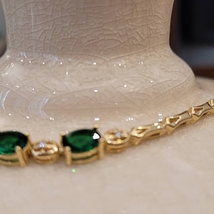 May include: A gold chain bracelet with emerald green oval-shaped gemstones. The bracelet is on a white surface.