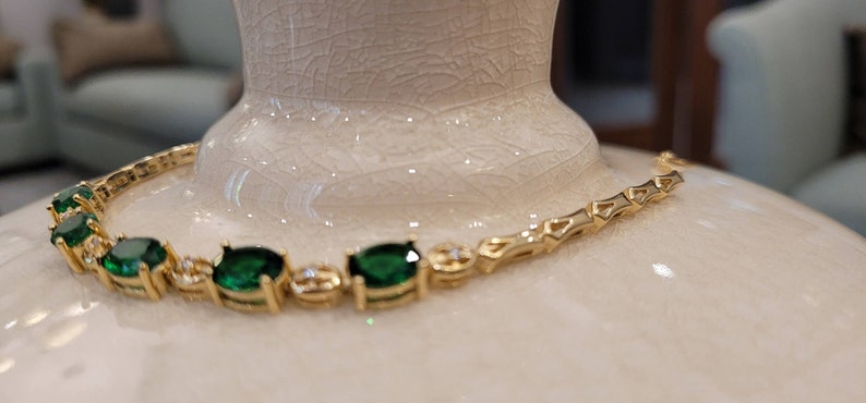 May include: A gold chain bracelet with emerald green oval-shaped gemstones. The bracelet is on a white surface.