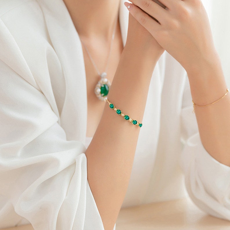 May include: A gold bracelet with a row of oval-shaped green gemstones. The bracelet is on a woman's wrist.