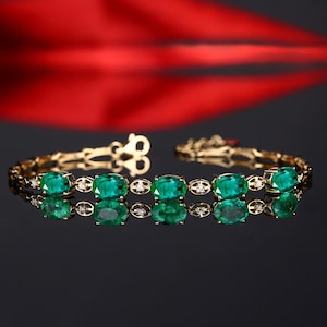 May include: A gold bracelet with a row of oval-shaped emerald gemstones and small diamonds set in between each emerald. The bracelet is reflected on a black surface.