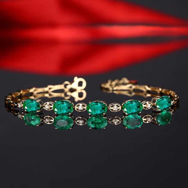 May include: A gold bracelet with a row of oval-shaped emerald gemstones and small diamonds set in between each emerald. The bracelet is reflected on a black surface.