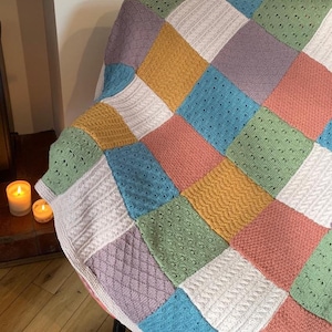 Peut inclure: Une couverture en patchwork tricotée avec des carrés de différentes couleurs, dont le blanc, le rose pâle, le bleu clair, le vert clair, le violet et le jaune. La couverture est posée sur une chaise.