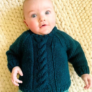 Puede incluir: Un bebé con un suéter de punto trenzado de color verde azulado y pantalones a rayas verdes y blancas. El suéter presenta un diseño de trenza central y puños acanalados. El bebé está acostado sobre una manta de punto grueso de color crema.