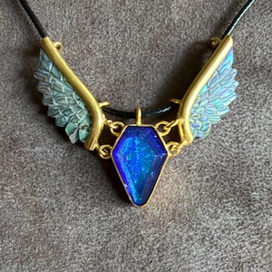 May include: A necklace featuring a central coffin-shaped pendant with a deep blue, glittery finish, framed in gold-tone metal. Two textured, wing-shaped accents in a muted teal-green hue extend from the sides, attached to a thin black cord.