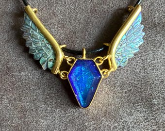 May include: A necklace featuring a central coffin-shaped pendant with a deep blue, glittery finish, framed in gold-tone metal. Two textured, wing-shaped accents in a muted teal-green hue extend from the sides, attached to a thin black cord.
