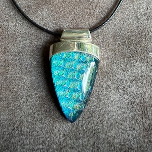 May include: A necklace featuring a triangular pendant with a textured, iridescent teal and blue glass inlay set in a polished silver-toned metal frame. The pendant hangs from a thin black cord secured with a silver-toned metal lobster claw clasp.