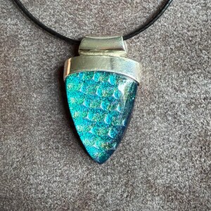 May include: A pendant necklace featuring a triangular piece of iridescent teal and blue dichroic glass with a textured scale pattern, set in a polished silver-toned metal cap and suspended from a simple black cord.