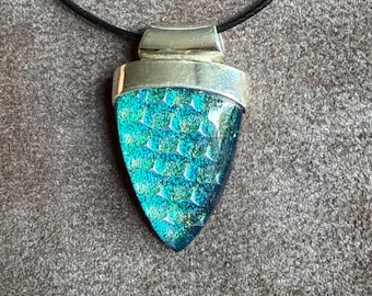 May include: A pendant necklace featuring a triangular piece of iridescent teal and blue dichroic glass with a textured scale pattern, set in a polished silver-toned metal cap and suspended from a simple black cord.