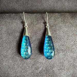 May include: A pair of drop earrings featuring teardrop-shaped pendants with a vibrant blue and green iridescent scale pattern. The pendants are suspended from silver-toned metal hooks, creating a sleek and reflective appearance for these jewelry pieces.