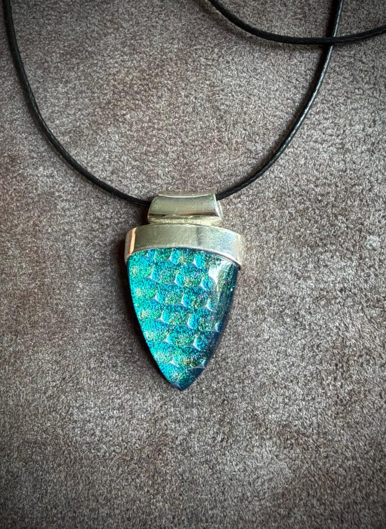 May include: A pendant necklace featuring a triangular piece of iridescent teal and blue dichroic glass with a textured scale pattern, set in a polished silver-toned metal cap and suspended from a simple black cord.