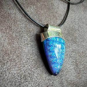 May include: A pendant featuring a teardrop-shaped piece of iridescent blue and purple glass with a textured, crystalline surface. The glass is set in a polished silver-toned metal cap and hangs from a simple black cord necklace.