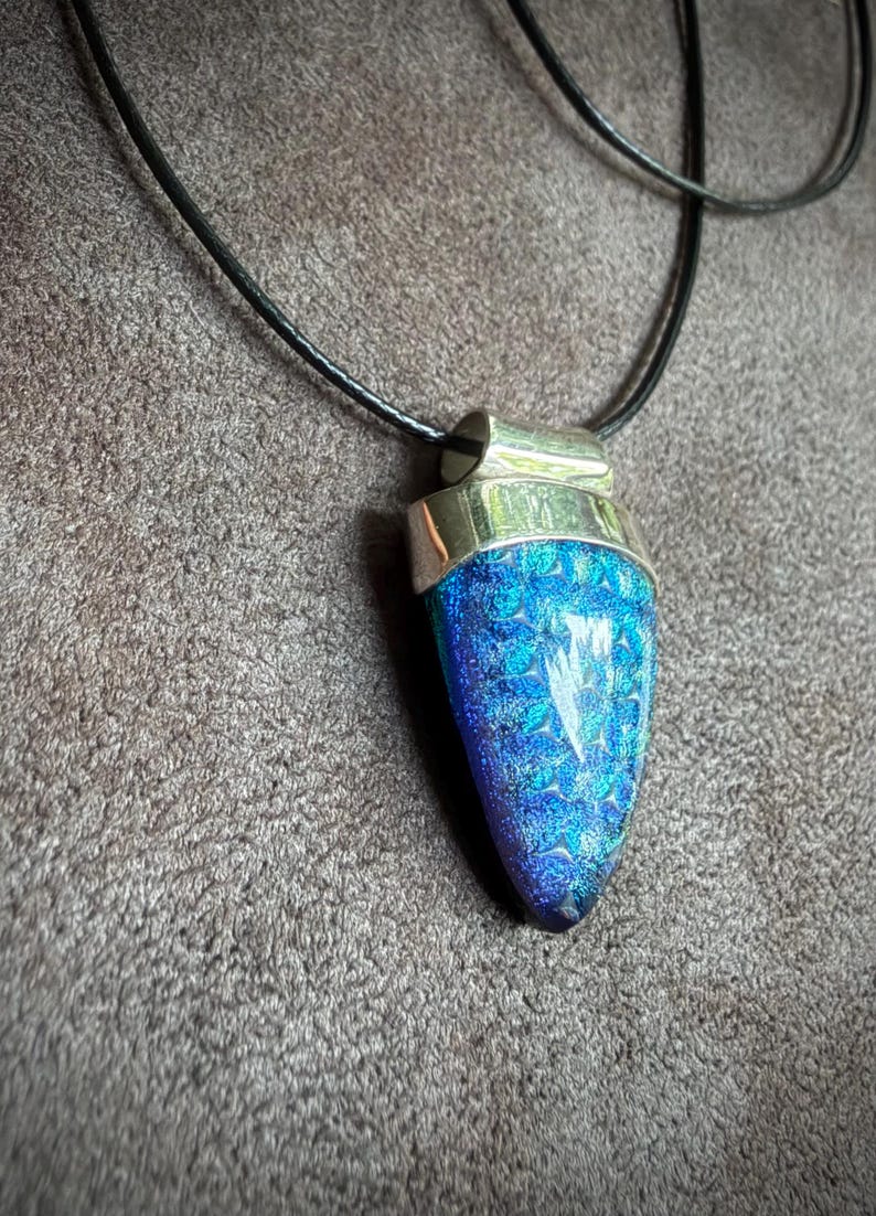 May include: A pendant featuring a teardrop-shaped piece of iridescent blue and purple glass with a textured, crystalline surface. The glass is set in a polished silver-toned metal cap and hangs from a simple black cord necklace.