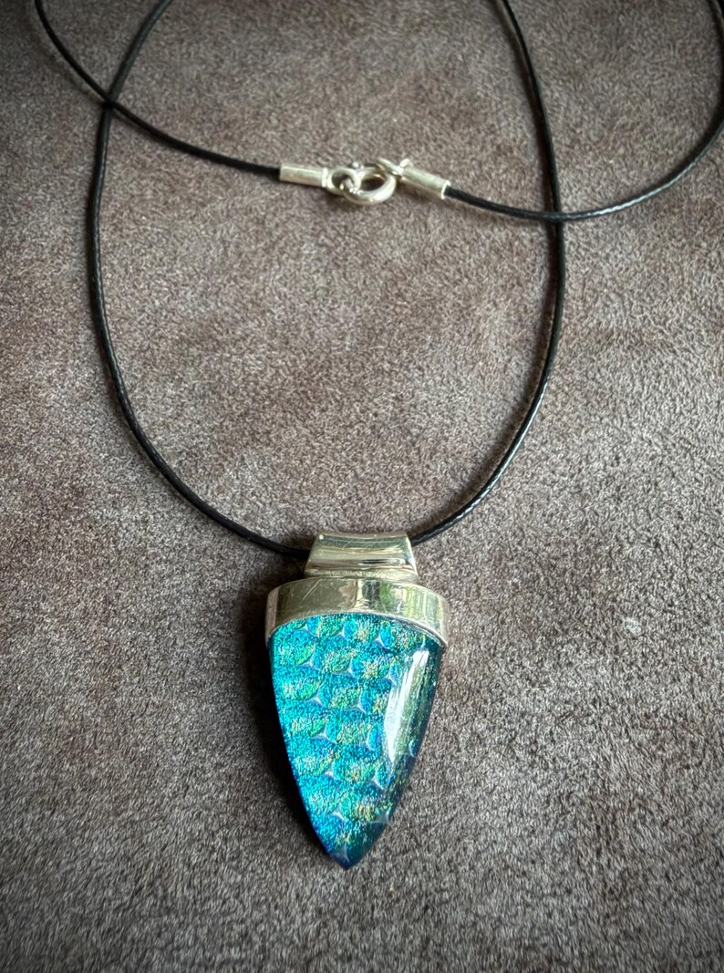 May include: A necklace featuring a triangular pendant with a textured, iridescent teal and blue glass inlay set in a polished silver-toned metal frame. The pendant hangs from a thin black cord secured with a silver-toned metal lobster claw clasp.