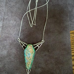 May include: A silver-toned necklace featuring a coffin-shaped pendant with an iridescent, multi-colored glass inlay. The pendant is held by a geometric metal frame with openwork detailing, suspended from a delicate silver-toned chain with a clasp.