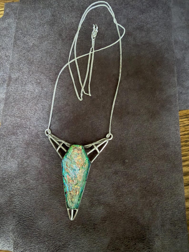 May include: A silver-toned necklace featuring a coffin-shaped pendant with an iridescent, multi-colored glass inlay. The pendant is held by a geometric metal frame with openwork detailing, suspended from a delicate silver-toned chain with a clasp.