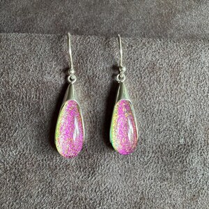 May include: A pair of drop earrings featuring teardrop-shaped glass cabochons with a vibrant pink and gold iridescent finish. The glass pieces are set in polished silver-tone metal caps and hang from matching silver-tone curved ear wires.