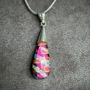 May include: A teardrop-shaped dichroic glass pendant featuring vibrant pink, orange, and teal geometric patterns. The glass is set in a polished silver-toned metal cap and hangs from a matching silver-toned chain necklace.