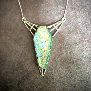 May include: A silver-toned necklace featuring a coffin-shaped pendant with an iridescent, multi-colored surface displaying shades of green, blue, and gold. The pendant is held by a geometric, open-work silver frame attached to a thin silver chain.