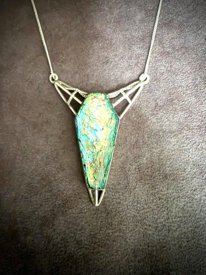 May include: A silver-toned necklace featuring a coffin-shaped pendant with an iridescent, multi-colored surface displaying shades of green, blue, and gold. The pendant is held by a geometric, open-work silver frame attached to a thin silver chain.