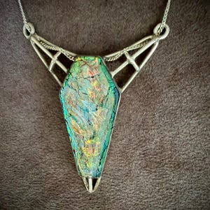 May include: A necklace featuring a coffin-shaped pendant with an iridescent, multi-colored glass surface displaying green, gold, and blue hues. The pendant is encased in a silver-toned metal frame with geometric, open-work detailing and a chain.