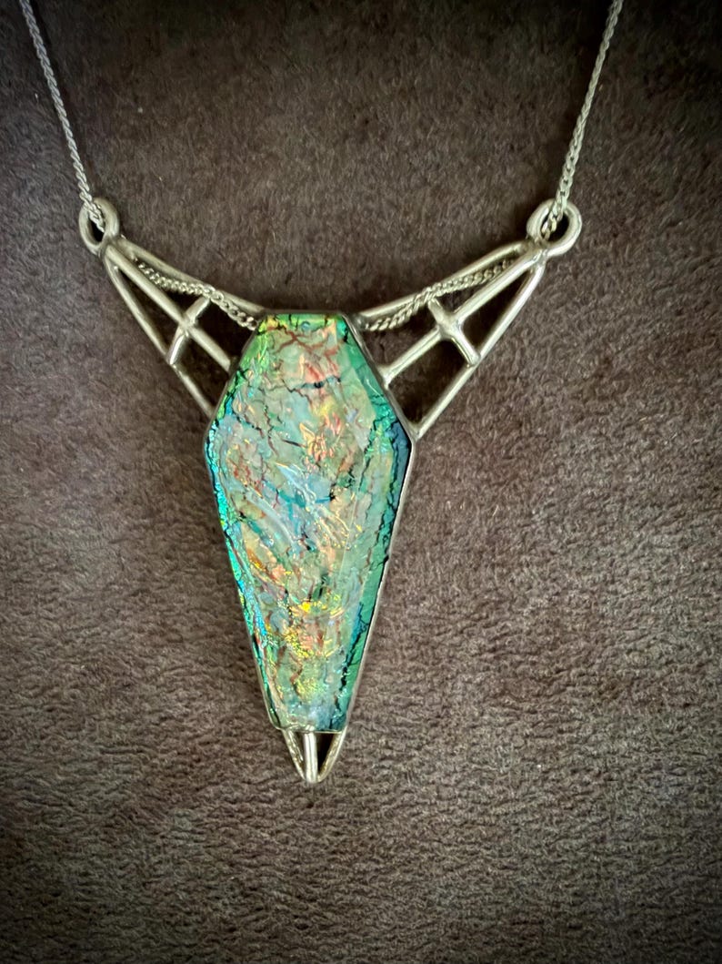 May include: A necklace featuring a coffin-shaped pendant with an iridescent, multi-colored glass surface displaying green, gold, and blue hues. The pendant is encased in a silver-toned metal frame with geometric, open-work detailing and a chain.