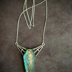 May include: A silver-toned necklace featuring a coffin-shaped pendant with an iridescent, multi-colored glass inlay. The pendant is held by a geometric metal frame connected to a thin silver-toned chain with a standard clasp closure.