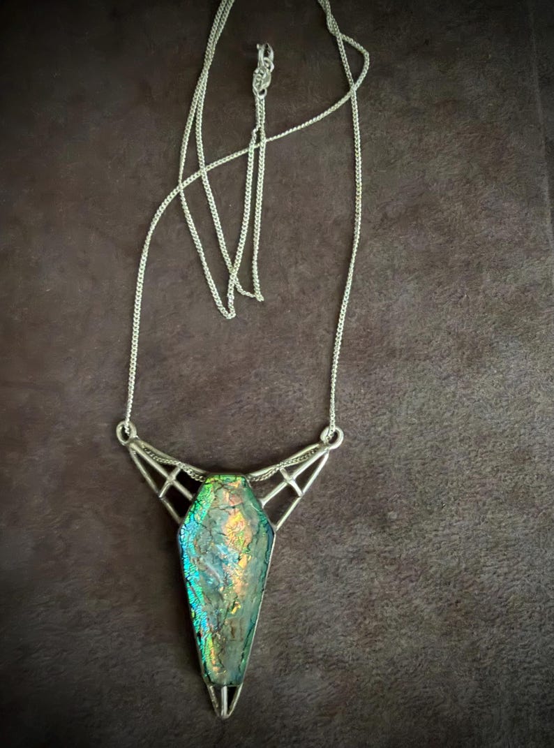 May include: A silver-toned necklace featuring a coffin-shaped pendant with an iridescent, multi-colored glass inlay. The pendant is held by a geometric metal frame connected to a thin silver-toned chain with a standard clasp closure.