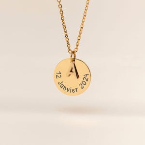 May include: A gold-coloured necklace with a circular pendant. The pendant features the letter "A" and the date "12 Janvier 2024" engraved around the edge. The chain is also gold-coloured and hangs from the pendant.