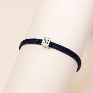 Op de afbeelding: Een marineblauwe armband met een zilverkleurige, kubusvormige bedel. De bedel heeft de letter "M" in een eenvoudig, schreefloos lettertype. De armband is gemaakt van twee strengen geweven koord.