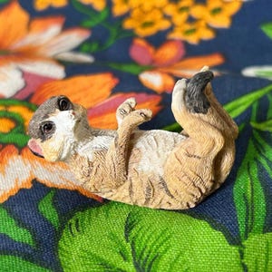 May include: A detailed figurine of a meerkat lying on its back, with its paws in the air. The meerkat has a light brown body with darker stripes and a grey head. The background is a colourful floral pattern.