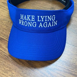 May include: A royal blue visor with a white embroidered message that reads "MAKE LYING WRONG AGAIN".