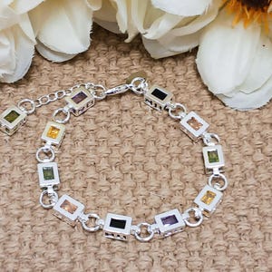 Puede incluir: Un brazalete de plata con una cadena y abalorios cuadrados, cada uno con una piedra preciosa de color diferente. Las piedras preciosas son moradas, verdes, amarillas, naranjas y negras.