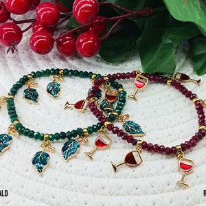 May include: Two beaded bracelets, one emerald green and one ruby red, adorned with gold charms. The emerald bracelet features leaf charms, while the ruby bracelet showcases wine glass charms. The bracelets are displayed with red berries and green leaves.