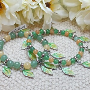 May include: Two green and yellow beaded bracelets with silver metal leaf charms. The bracelets are on a white and green textured surface. The text "aventurine" is visible on a white tag.