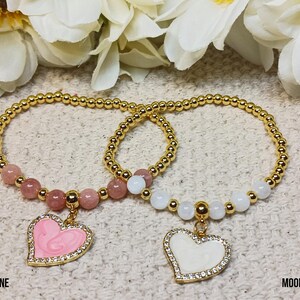 May include: Two gold bracelets with heart charms. One bracelet has pink and white beads and a pink heart charm. The other bracelet has white beads and a white heart charm. The bracelets are labeled "Sunstone" and "Moonstone".