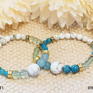 May include: Two bracelets with white, light blue, dark blue, and green beads. The bracelets have gold accents and are on a beige surface. The text "STYLE 1" and "STYLE 2" are visible below the bracelets.