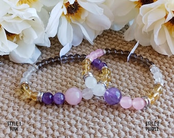 May include: Two beaded bracelets with semi-precious stones. One bracelet features pink and purple stones, while the other has yellow and clear beads. The bracelets are displayed on a textured surface with white flowers in the background. Style 1: Pink, Style 2: Purple.