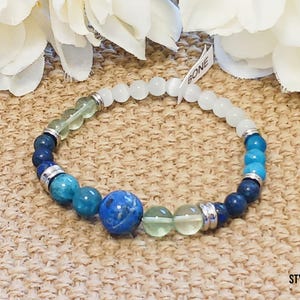 Pode incluir: Um bracelete de contas azuis, verdes e brancas com detalhes prateados. O bracelete tem uma conta azul grande no centro. O texto "BONE" &eacute; vis&iacute;vel em uma etiqueta branca.