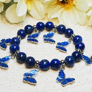 以下が含まれることがあります： 青いラピスラズリビーズのブレスレットに、銀色の蝶のチャームが飾られています。ブレスレットは丸いビーズで作られており、伸縮性のあるゴムバンドが付いています。