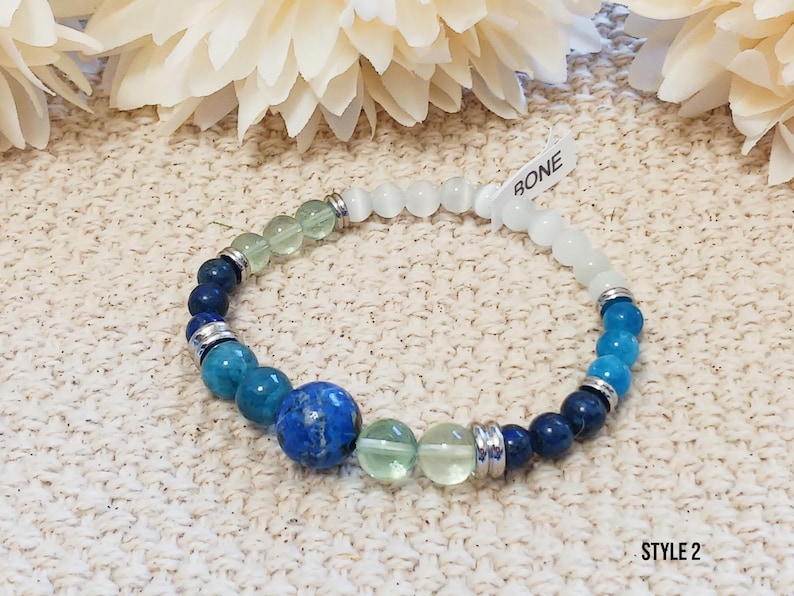 Pode incluir: Um bracelete de contas azuis, verdes e brancas com um fecho de prata. O bracelete tem uma etiqueta que diz "BONE" e "STYLE 2".